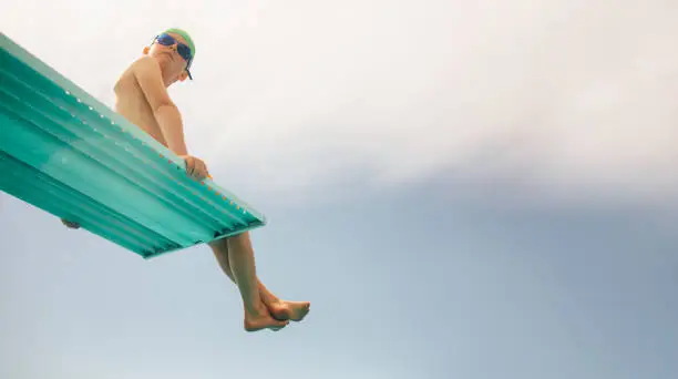 Low angle view of boy in swim goggles and swimming cap sitting on diving board against bright sky. Boy on diving platform at pool.