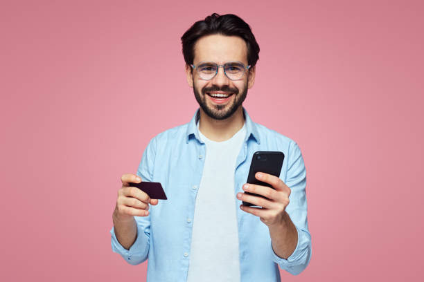 istock Attractive handsome man smiling while holding plastic credit card and mobile phone, isolated over pink background. 1142850863