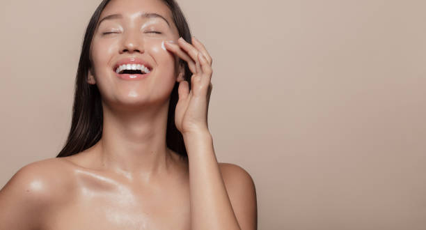 Happy with her beauty regime Beautiful girl with bare shoulders applying cream on her face and smiling against beige background. Smiling asian woman with glowing skin applying facial skincare cream with eyes closed. moisturizer photos stock pictures, royalty-free photos & images