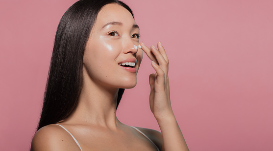 Close up of a youthful female model applying moisturizer to her face. Young korean woman putting moisturizer cream on her pretty face against pink background.