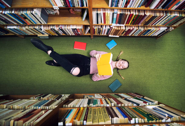 joven estudiante acostado en el piso de la biblioteca y libro de lectura. - bookshelf learning education study fotografías e imágenes de stock