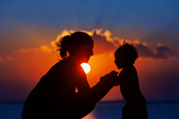 Black silhouette of mother, baby son walk by sea beach Happy family black silhouette on sunset sky background. Young mother, baby son have fun together, walk by sea beach. Barefoot child look at sun. Travel lifestyle, parents with kids on summer vacation. raro stock pictures, royalty-free photos & images