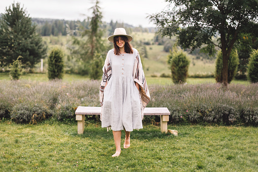 Stylish hipster girl in linen dress and hat walking at lavender field and relaxing in mountains. Bohemian woman smiling and enjoying vacation. Atmospheric rustic moment. Copy space