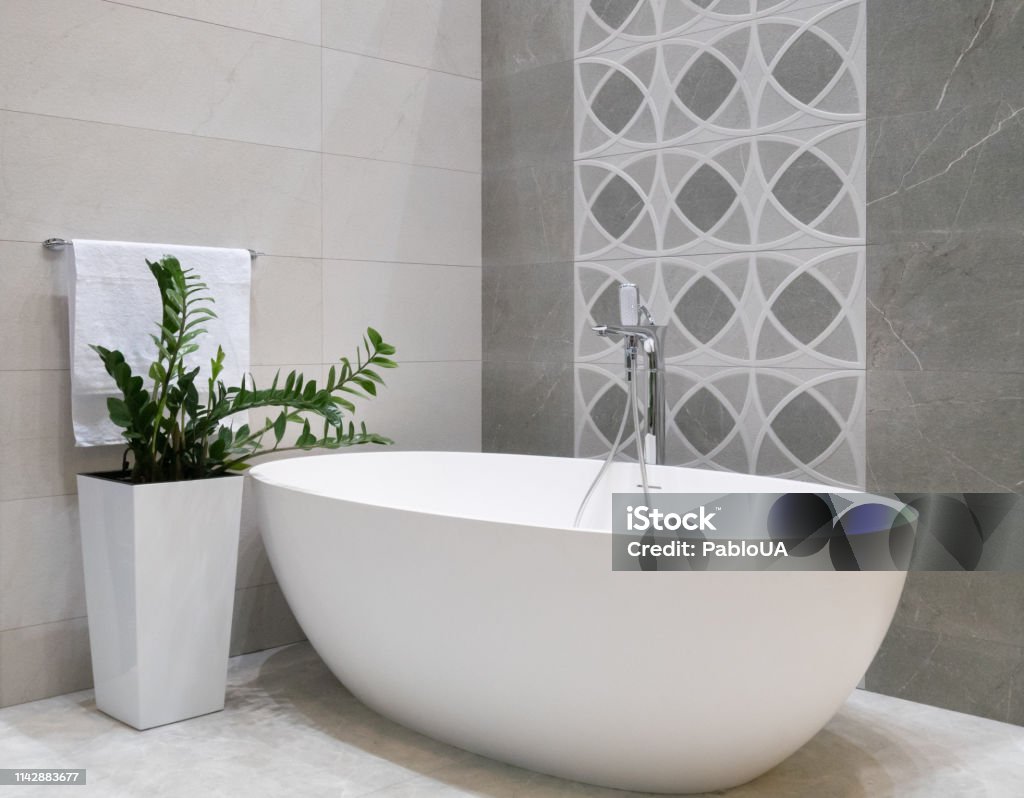 modern bathroom interior design with white stone bathtub, grey tiles wall, ceramic flowerpot with green plant and hanger with towel Bathroom Stock Photo