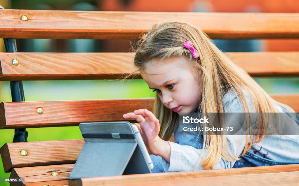 Cute little girl having fun with tablet in the park, watching cartoon. Lifestyle, technology concept Cute little girl having fun with tablet in the park, playing video games. Lifestyle, technology concept Applying Stock Photo