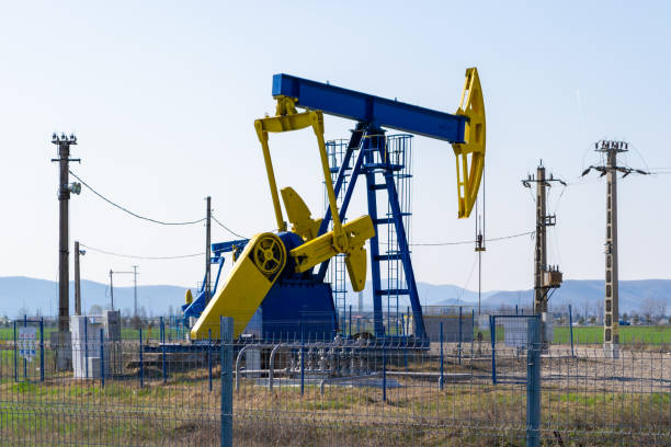 синий и желтый насосный домкрат над нефтяной колодец, окруженный столбами электричества и защищенный забором, при ярком дневном свете, лет� - omv стоковые фото и изображения