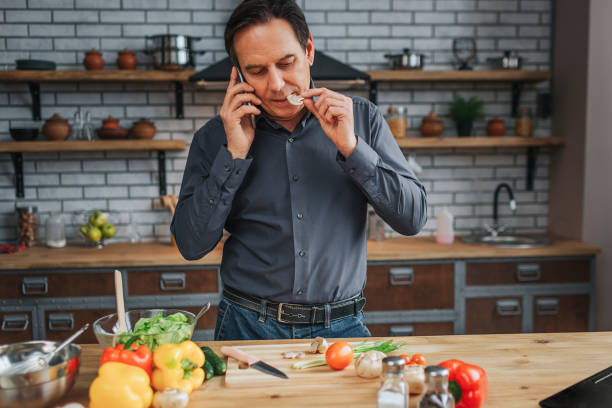 erwachsener mann steht am küchentisch und telefoniert er schmeckt stück pilz. man schaut nach unten. buntes gemüse, das auf dem schreibtisch liegt. - smart casual occupation casual healthy eating stock-fotos und bilder