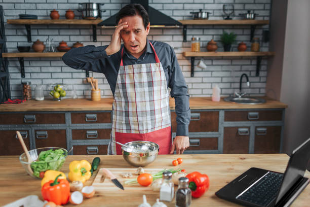 istock Scared man stant at table in kitchen and look at it. He hold hand on head. Man wear apron. Colorful vegetables lying on desk. 1142910901