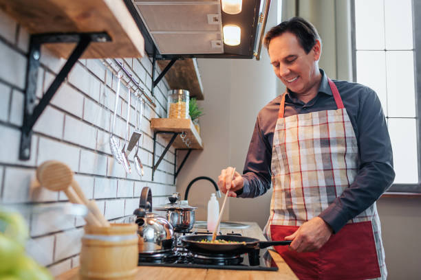 fröhlicher mann in schürze stehen am herd und kochen essen. er lächelt und mischt zutaten. der mensch steht allein da. - smart casual occupation casual healthy eating stock-fotos und bilder