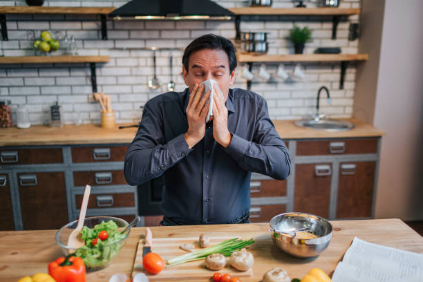 Sick adult man sneezing to white napkin. He stand at table in kitchen. Desk full of colorful healthy vegetables and spices. stock photo