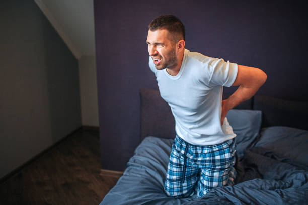 istock Young man is screaming. He holds hands on back and look forward. He wears pajamas. Guy is suffering. Pain is strong. He is in bedroom. 1142911743