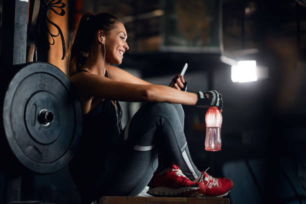 Happy sportswoman using cell phone on water break in a gym. Young smiling athletic woman relaxing on water break and reading text message on her smart phone in a gym. groyne photos stock pictures, royalty-free photos & images