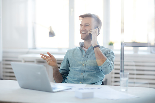 Portrait of handsome young man laughing cheerfully while speaking by phone sitting at desk in office, copy space