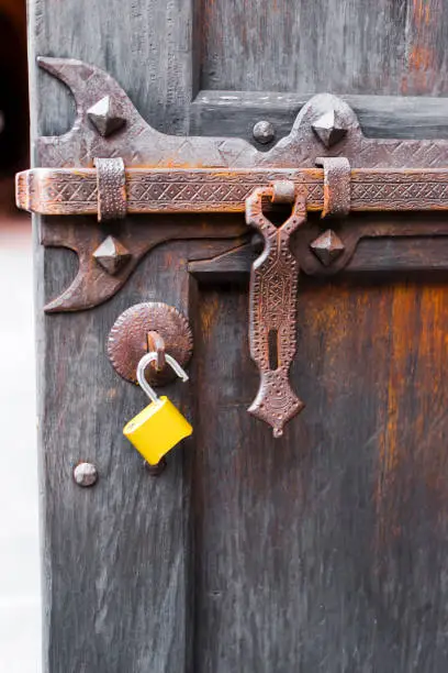 Powerful Forged bolt carved with ornaments and parts for closure on an old wooden door with rust on the deadbolt and modern open yellow padlock, as a meeting of old and new eras.