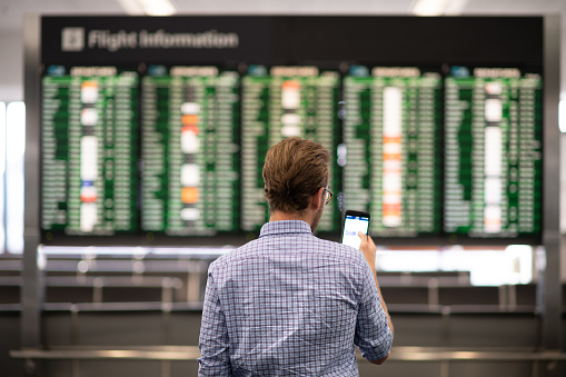Businessman references the flight information board to check if his flight will be on time. He holds his smartphone up to compare the times.