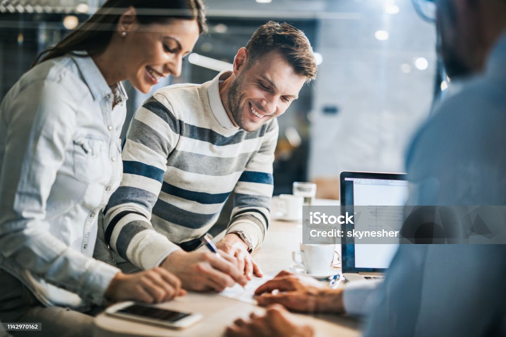 Happy couple signing a contract on a business meeting with their insurance agent. Young happy couple signing a document while being on a meeting with their real estate agent in the office. The view is through glass. Banking Stock Photo