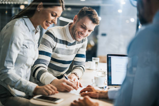 Happy couple signing a contract on a business meeting with their insurance agent. Young happy couple signing a document while being on a meeting with their real estate agent in the office. The view is through glass. mortgage document stock pictures, royalty-free photos & images