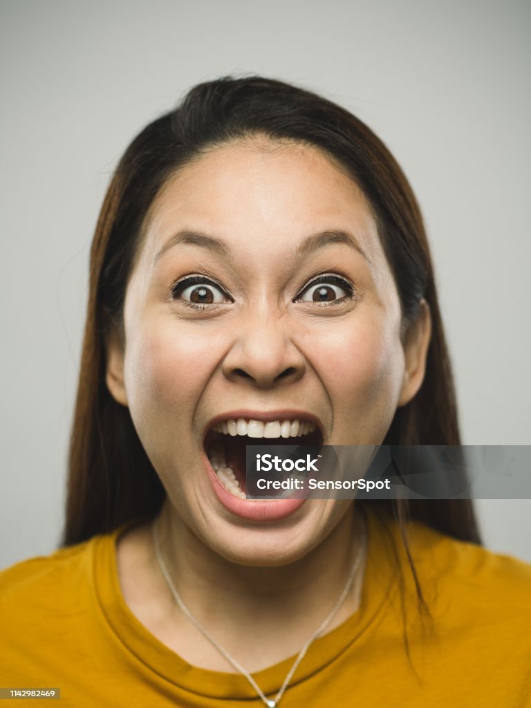 Portrait of real malaysian young woman with shouting expression Close up portrait of asian young woman with shouting expression against  white gray background. Vertical shot of malaysian real people screaming in studio with long brown hair. Photography from a DSLR camera. Sharp focus on eyes. Women Stock Photo