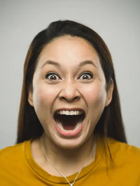 Close up portrait of asian young woman with shouting expression against  white gray background. Vertical shot of malaysian real people screaming in studio with long brown hair. Photography from a DSLR camera. Sharp focus on eyes.