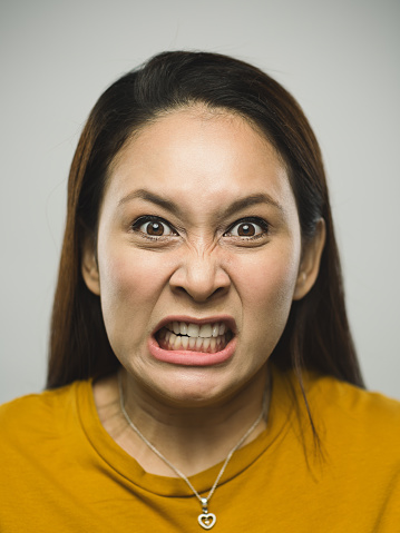 Close up portrait of asian young woman with very angry expression against white gray background. Vertical shot of malaysian real people furious in studio with long brown hair. Photography from a DSLR camera. Sharp focus on eyes.
