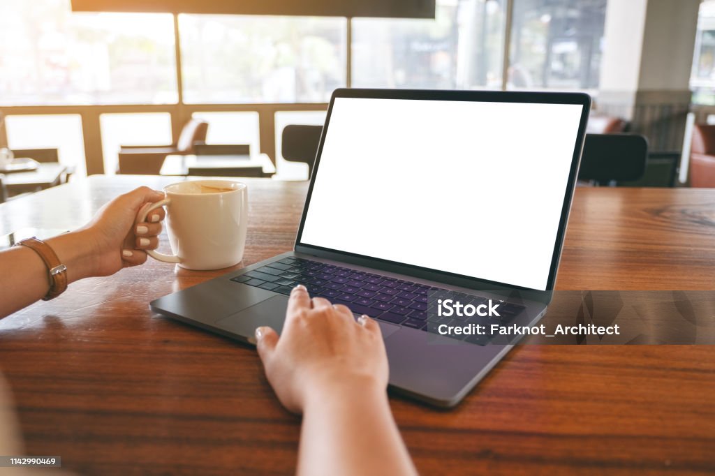 a woman using and touching on laptop touchpad with blank white desktop screen on wooden table while drinking coffee Mockup image of a woman using and touching on laptop touchpad with blank white desktop screen on wooden table while drinking coffee Above Stock Photo