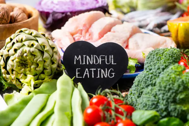 a black heart-shaped signboard with the text mindful eating, on a pile of different vegetables, such as French beans, cherry tomatoes, a head of broccoli, and some pieces of chicken in the background