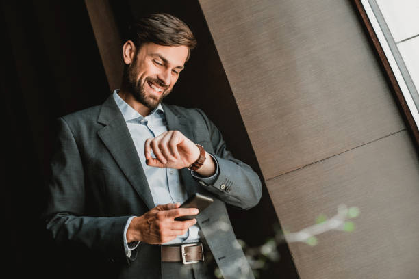 Cheerful CEO is monitoring time on watch Low angle of smiling businessman wearing suit and standing indoor. He is holding mobile phone and checking time on wrist checking the time stock pictures, royalty-free photos & images