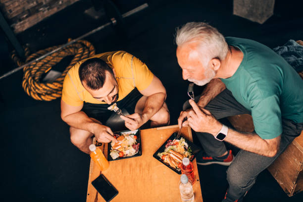 Active senior sportsman eating a healthy protein salad meal in a take-out box with his personal trainer in a gym area of a gym