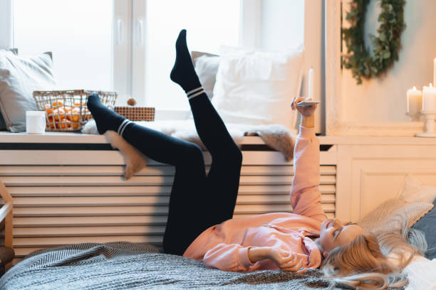 コケット若い女の子は、窓の近くに、自宅でベッドの上に横たわっている間、自分撮りを作り、楽しみを持って、 - furniture internet adult blond hair ストックフォトと画像