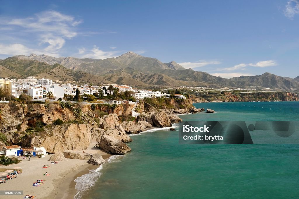 Rugged coastline in Nerja in Malaga, Spain The view of the Nerja in Malaga, Spain, is pictured.  There are mountains and an outcropping of rock to the left of the sea dotted with white and pale-colored houses.  The sea is teal and blue with a blue sky and wispy white clouds in the background. Andalusia Stock Photo