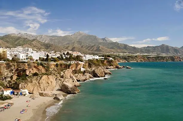 The view of the Nerja in Malaga, Spain, is pictured.  There are mountains and an outcropping of rock to the left of the sea dotted with white and pale-colored houses.  The sea is teal and blue with a blue sky and wispy white clouds in the background.
