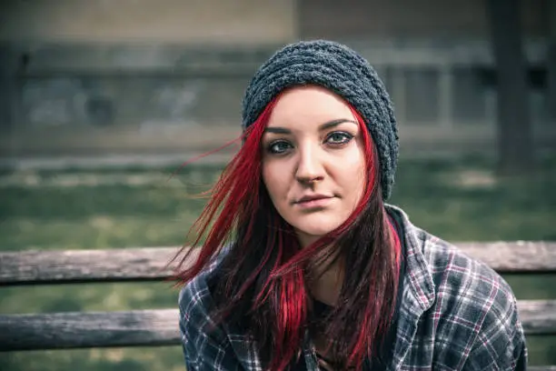 Photo of Young beautiful red hair girl sitting alone outdoors on the wooden bench with hat and shirt feeling anxious and depressed after she became a homeless person close up portrait