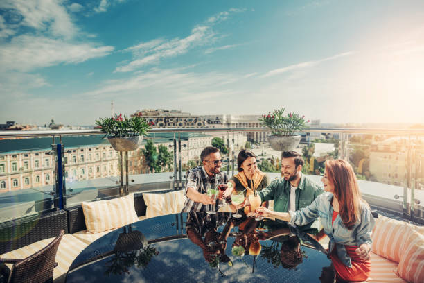 Cheerful young people enjoying drinks at rooftop bar Time to celebrate. Smiling friends raising cocktails for toasting. They are sitting at the table with their mirror reflection restaurant mirror stock pictures, royalty-free photos & images