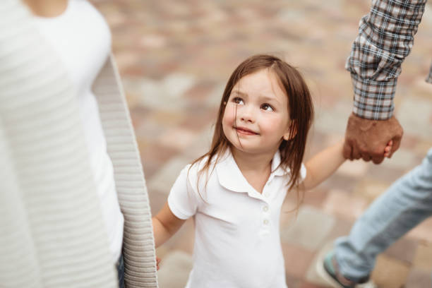 Small girl enjoying time with parents Waist up portrait of little kid looking at mom with joy. She is walking with mother and father holding hands and feeling delighted kid holding fathers hand stock pictures, royalty-free photos & images