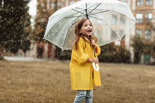 Waist up portrait of joyful kid spending time outside standing with umbrella in hands. She is looking upwards with content and sincere smile