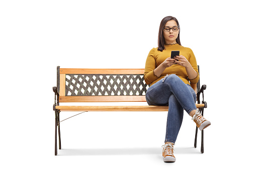 Young female sitting on a bench and typing on a mobile phone isolated on white background