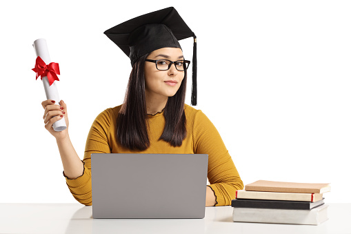 Young woman with a graduation hat and diploma sitting with a laptop and books isolated on white background