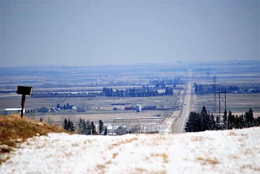 A country rural scene of a long road with beautiful landscape and scenery of the valley below. taken after a recent snowfall, snow remains on the ground next to a rural mail box on the left side, some evergreens can be seen in the distance.