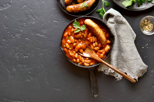 Sausages with baked white beans in tomato sauce White beans in tomato sauce and grilled sausages in frying pan over black stone background with copy space. Top view, flat lay baked beans stock pictures, royalty-free photos & images