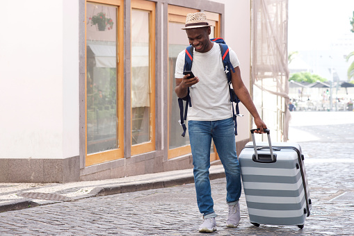 Full body portrait of happy african american tourist walking with suitcase and mobile phone