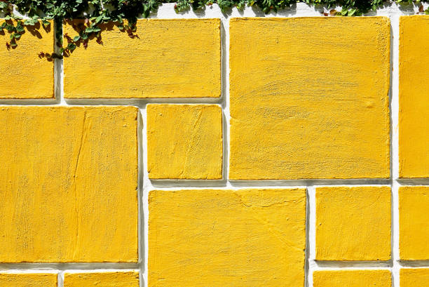 Detail of a rectangle shaped grid design of a concrete garden wall fence painted in bright yellow. Green plants hanging from the top of the wall.