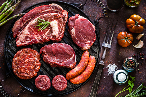 Various cuts of raw meat shot from above on rustic kitchen table. The cuts are on a cast iron grill and includes Angus steak, tenderloin, sausages and hamburger meat. The grill is surrounded by herbs, vegetables and spices for cooking meat like peppercorns, salt, garlic, tomatoes, asparagus and rosemary. A vintage fork is included in the composition. Predominant colors are red and brown. Low key DSRL studio photo taken with Canon EOS 5D Mk II and Canon EF 100mm f/2.8L Macro IS USM.