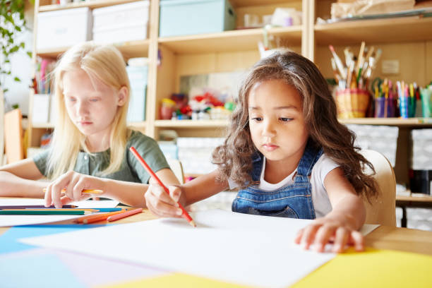 istock Two lovely girls sitting at table and drawing with colored pencils while attending art class in school 1143087264