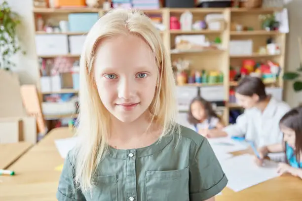 Photo of Lovely albino girl smiling and looking at camera while standing on blurred background of classroom in art school