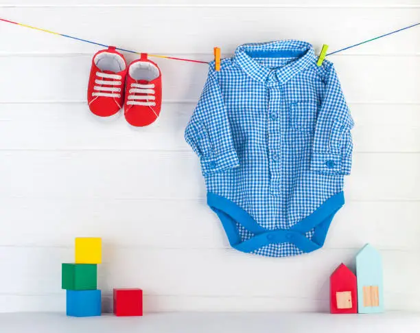Red baby booties and blue bodykit on clothespins and wooden toys over white wooden background with blank space for text.
