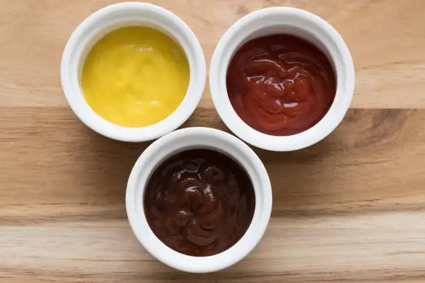 Closeup of three mini ramekin with ketchup, mustard sauce and barbecue sauce on wooden background.