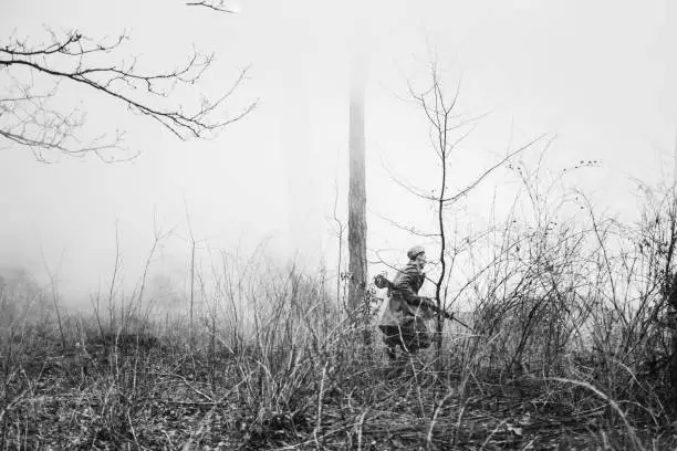 Re-enactor Dressed In Overcoat As World War II Russian Soviet Red Army Soldier Running In Fog Smoke With Machine Gun In Misty Forest Ground. Photo In Black And White Colors. Soldier Of WWII WW2 Times
