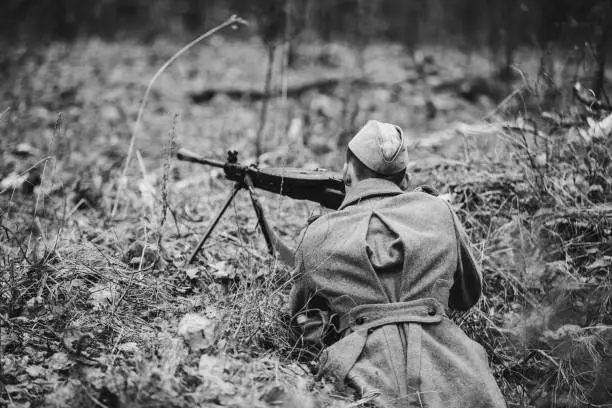 Re-enactor Dressed In Overcoat As World War II Russian Soviet Red Army Soldiers Hidden Aiming With Machine Gun In Forest Ground. Photo In Black And White Colors. Soldier Of WWII WW2 Times.