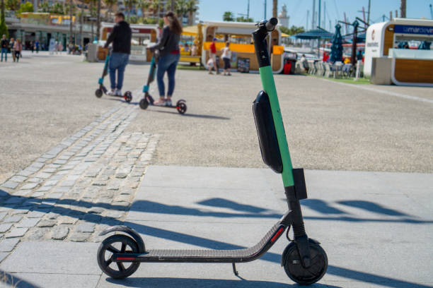 electric scooter roller parked on the sidewalk at the port of Malaga on a sunny day and a blurred couple use them in background modern transportation system Spain, Malaga - 04.04.2019: electric scooter roller parked on the sidewalk at the port of Malaga on a sunny day and a blurred couple use them in background modern new transportation system. lime scooter stock pictures, royalty-free photos & images