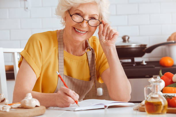 Grandma cooking at the morning, writing something in recipe book Grandma in eyeglasses cooking at the morning, writing something in recipe book, looking at camera. Indoor, studio shoot, kitchen interior finely stock pictures, royalty-free photos & images